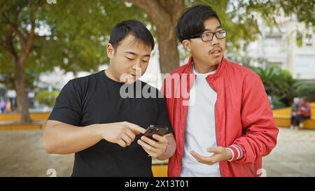 Two asian men browsing a smartphone together in a lush city park. Stock Photo