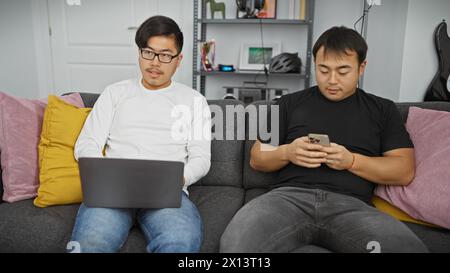 Two asian men relax on a sofa, one using a laptop and the other a smartphone, in a modern living room. Stock Photo