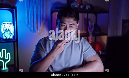 A young asian man sending a voice message indoors at night in a room with neon lighting. Stock Photo