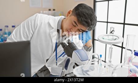 A young asian man in a lab coat appears exhausted while working with a microscope in a laboratory setting. Stock Photo