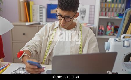A young man with measuring tape is focused on his mobile phone in a colorful tailor shop with sewing machine and fabric Stock Photo