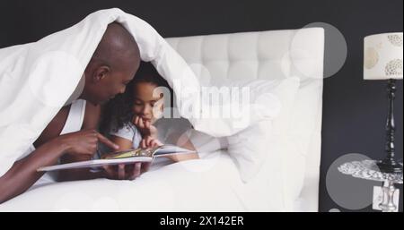 Image of light spots and trails over african american man and his daughter reading book together Stock Photo