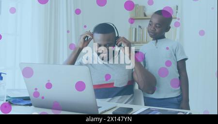 Image of pink spots floating over african american father and son using phone headset and laptop Stock Photo