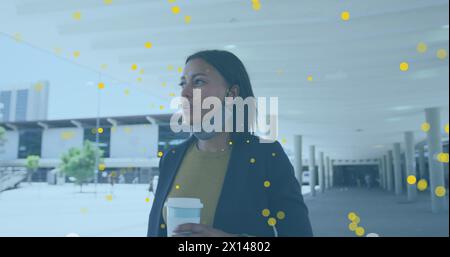Image of yellow spots floating over african american woman holding coffee cup walking at office Stock Photo