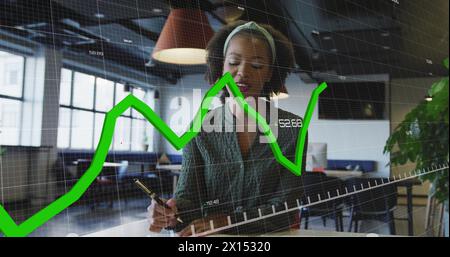 Image of graphs and changing numbers over biracial woman writing in notepad Stock Photo
