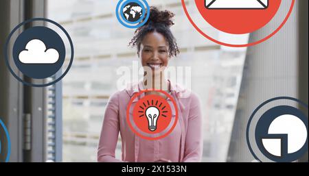Image of icons over smiling biracial woman standing arms crossed against glass window Stock Photo