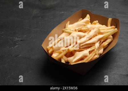 Tasty potato fries and cheese sauce in paper container on black table, closeup Stock Photo