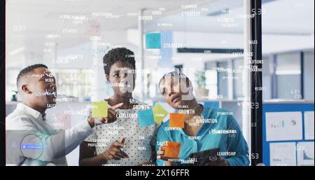Image of business words and numbers over diverse colleagues discussing over notes on glass wall Stock Photo