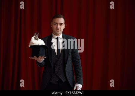 Young handsome magician holding top hat with cute fluffy rabbit Stock Photo