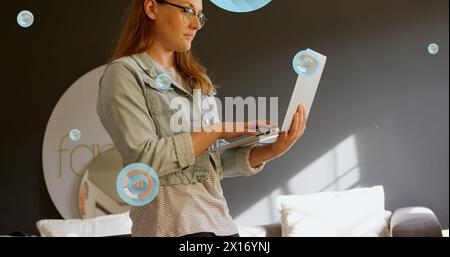 Image of multiple graph icons over caucasian woman standing and using laptop in office Stock Photo