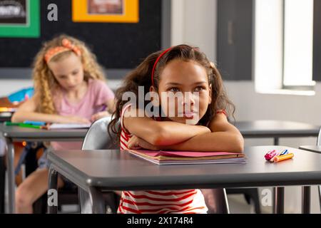 In school, young biracial female student sitting at a desk in a classroom, looking thoughtful Stock Photo