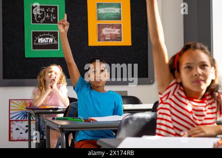 In school, diverse group of young students sitting at desks in a classroom, raising hands Stock Photo