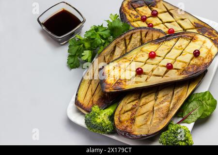 Baked eggplant with with parsley and broccoli. Sauce in bowl and cranberries on table. Gray background. Top view Stock Photo