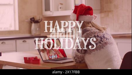 Image of happy holidays text over happy caucasian woman in santa hat on laptop image call Stock Photo