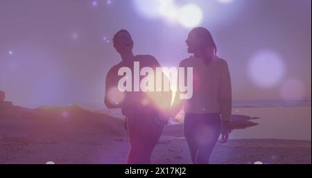 Image of glowing spots of light over african american couple walking on the rocks near the sea Stock Photo
