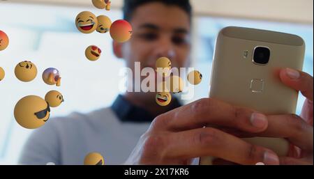 Image of multiple face emojis floating over biracial man using smartphone at office Stock Photo