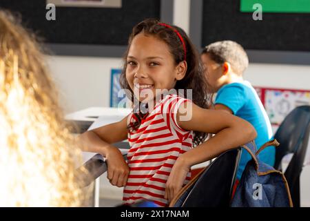 In school, three children wearing casual clothes are sitting at desks in a classroom. One girl in a striped shirt is turning around, smiling at someon Stock Photo