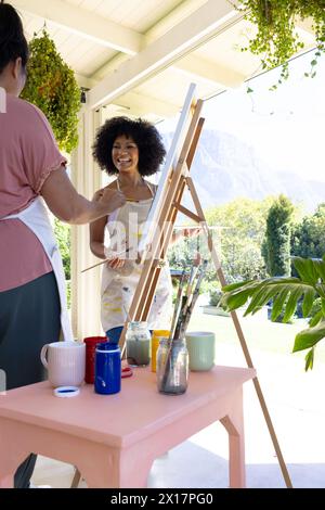 Mature biracial woman painting at home, young biracial woman watching, both smiling Stock Photo