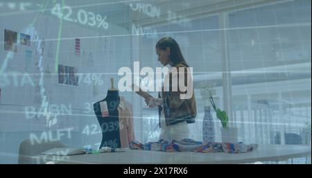 Image of graphs and trading board on biracial female designer examining design of dress Stock Photo