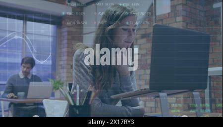 Image of trading board and graphs over caucasian woman working on laptop at office Stock Photo