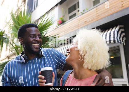 Diverse couple laughing, holding coffee outside cafe Stock Photo