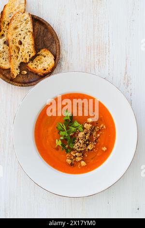 Squash soup with bread crumbs, parsley, and chili oil in plate served with crusty bread on white wood background, high angle view Stock Photo