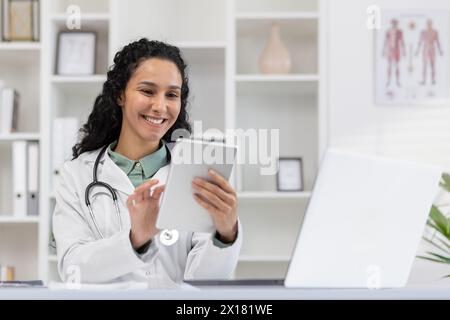 Cheerful female doctor in a white coat delights in using a digital tablet in her bright medical office, exemplifying professional happiness and digital healthcare. Stock Photo
