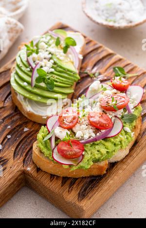 Avocado toasts on a wooden board with cream cheese, radishes and feta topped with red onion and freshly ground pepper Stock Photo