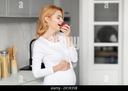 Expectant mother eating apple in kitchen, copy space Stock Photo