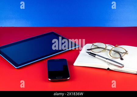 Mobile phone with tablet device and notebook pen and eye glasses on a red base and blue background Stock Photo