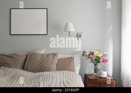 Elegant bedroom. Tulips, cherry tree blossoms bouquet in glass vase. Wooden night stand. Cup of coffee. Blank black picture frame mockup. Lamp Stock Photo