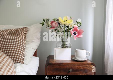 Spring interior still life. Blank greeting card, invitation mockup. Elegant bedroom. Tulips, cherry tree blossoms bouquet in glass vase. Wooden night Stock Photo
