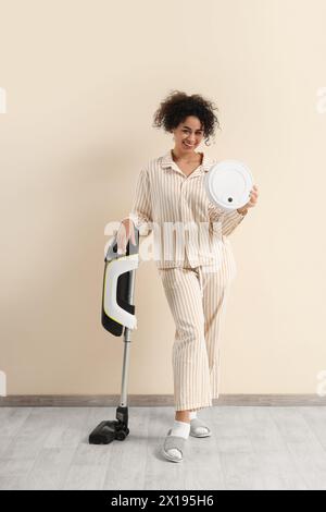Young African-American woman in pajamas with vacuum cleaners near beige wall Stock Photo