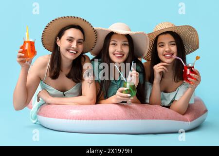 Beautiful young women in wicker hats with glasses of cocktail lying on inflatable mattress against blue background. Summer party Stock Photo