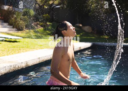 A teenage Asian boy standing in a pool outside at home, splashing water on his face Stock Photo