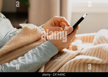 a caucasian man relaxing, using smart phone lying in bed under throw at home Stock Photo