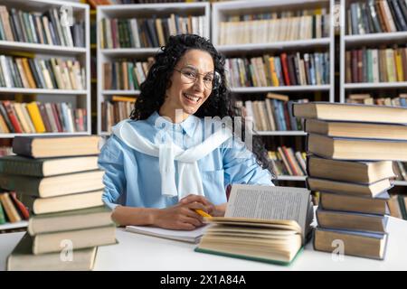 A cheerful young woman with glasses and curly hair studies among stacks of books in a library, exuding curiosity and enthusiasm. Stock Photo