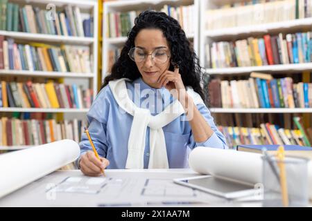 A professional female architect deeply focused on designing architectural blueprints at a library desk, surrounded by books. Stock Photo