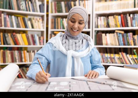 A cheerful Muslim woman architect drawing and reviewing building plans at a table in a university library. She's dressed in a hijab. Stock Photo