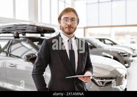 Young successful manager on background of rows of new car dealership crossovers. Portrait of confident Caucasian man, salesman in suit in bright moder Stock Photo