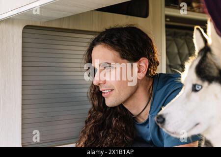 Horizontal photo a smiling man with curly hair and his attentive husky companion look out together from the comfort of their camper van, cap Stock Photo