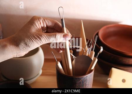Woman taking clay crafting tool from cup in workshop, closeup Stock Photo