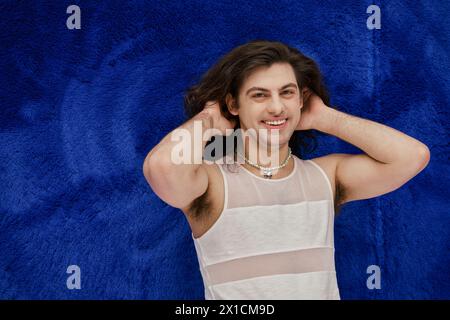 handsome cheerful gay man with long hair smiling at camera on dark blue backdrop, pride month Stock Photo