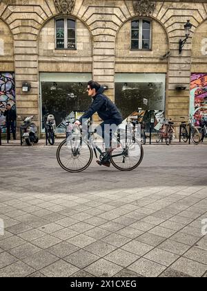 Bordeaux, France -- April 16, 2024. A young man dressed in black and wearing sunglasses, pedals a bike down a Bordeaux street in France. Stock Photo