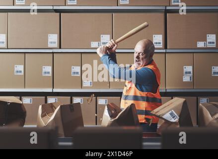 Angry rebellious worker smashing boxes with a bat on the conveyor belt, he is frustrated and furious Stock Photo