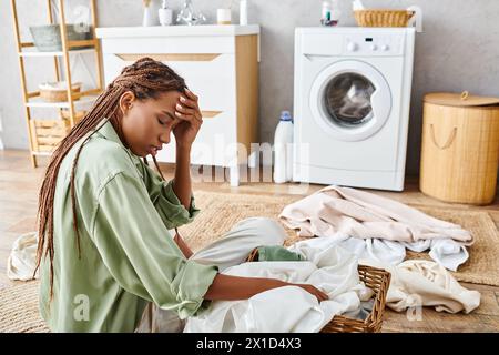 A woman with afro braids sits next to a pile of clothes, lost in thought while doing laundry in a bathroom. Stock Photo