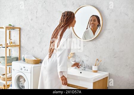 African American woman in bathrobe with afro braids stands in front of modern washer in stylish bathroom. Stock Photo