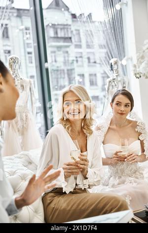 A young bride in a wedding dress, her bridesmaid, and a middle-aged mother sitting closely in a bridal salon. Stock Photo