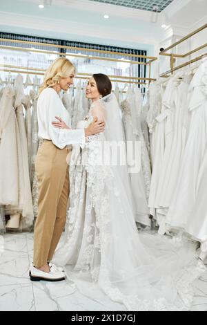 A young brunette bride in a wedding dress and her middle-aged mother stand side by side in a bridal salon, looking at a rack of gowns. Stock Photo