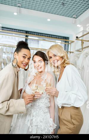 A young bride in a wedding dress stands between her middle-aged mother and best friend in a bridal salon, smiling warmly. Stock Photo
